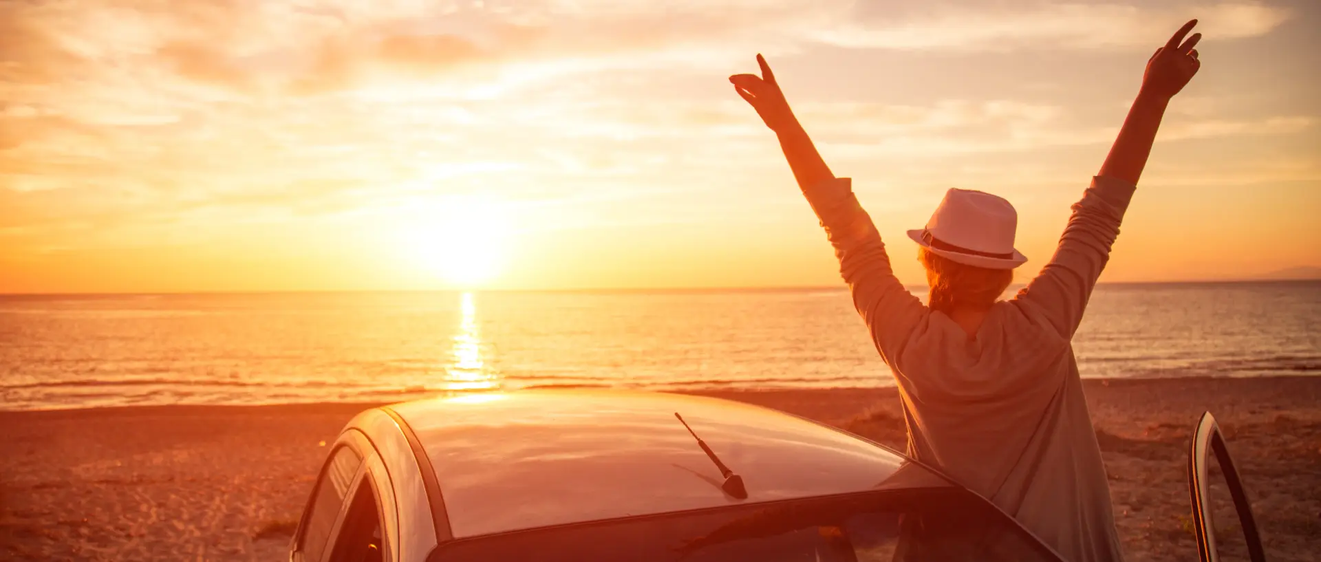 Frau mit Auto steht am Meer und geniesst den Sonnenuntergang