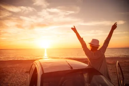 Frau mit Auto steht am Meer und geniesst den Sonnenuntergang